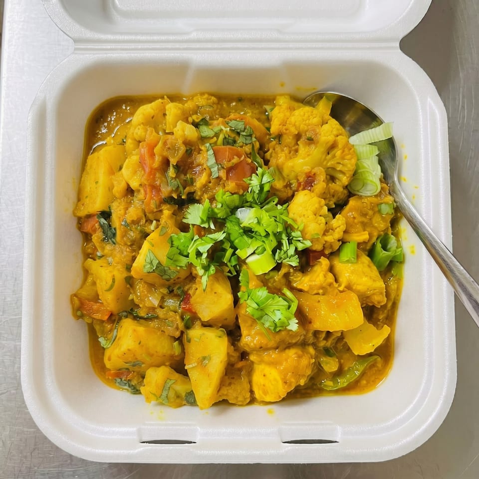 Aloo Gobi, a vibrant Indian dish with potatoes and cauliflower, garnished with fresh cilantro in a takeout container.