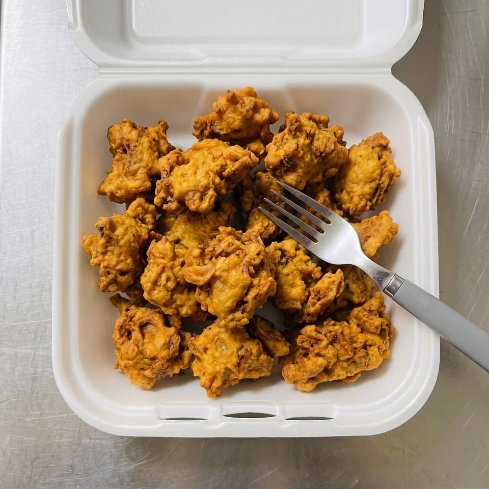 Golden-brown fried pakora in a white takeout container with a fork on a metal surface.