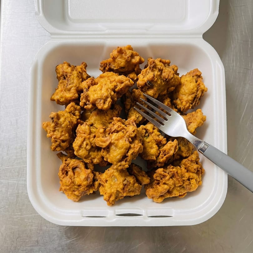 Golden-brown fried pakora in a white takeout container with a fork on a metal surface.