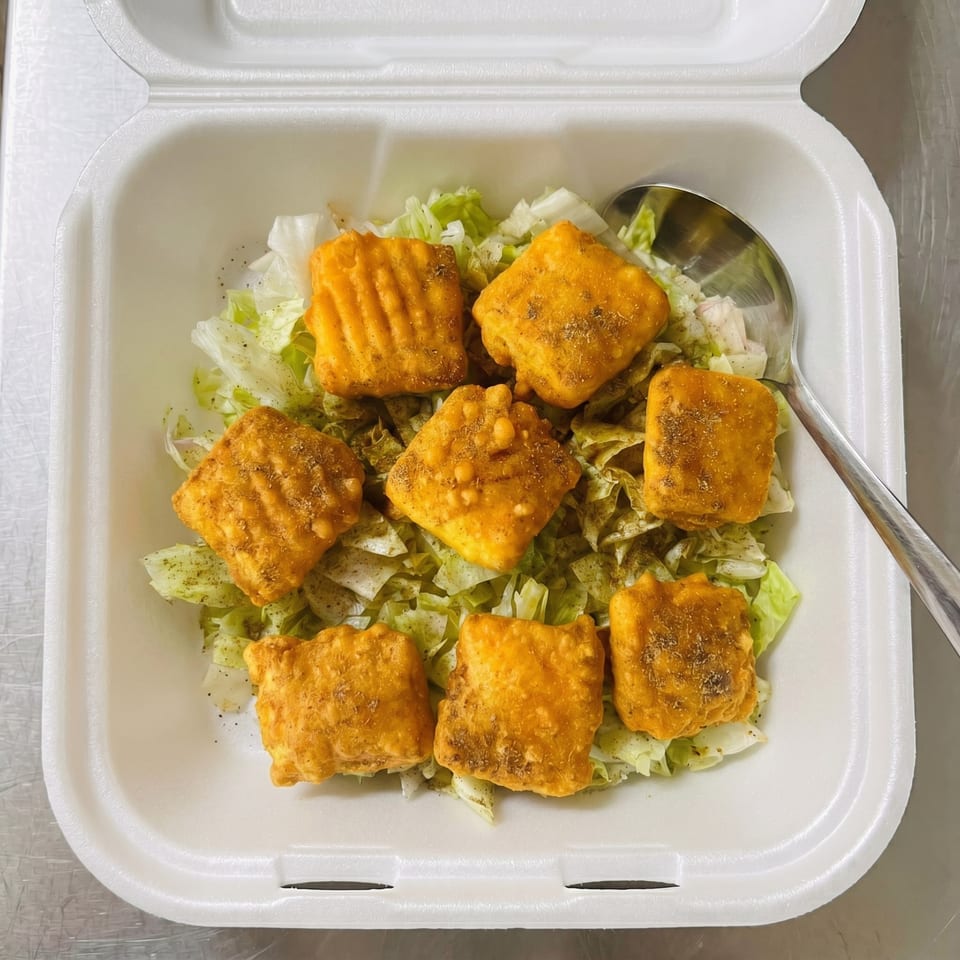 Seven golden-brown fried paneer pieces with seasoning on a bed of shredded lettuce in a white takeout container.