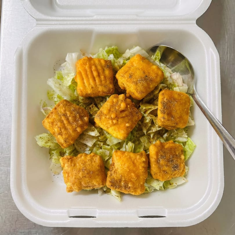 Seven golden-brown fried paneer pieces with seasoning on a bed of shredded lettuce in a white takeout container.